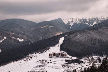 A snow-covered mountainous landscape with dense forests and a ski resort nestled in the valley. The resort consists of several buildings, surrounded by winding roads. In the distance, snow-capped peaks rise under a cloudy sky.