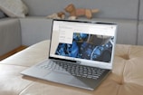 A sleek ASUS laptop is placed on a beige ottoman. The screen displays a webpage featuring a design with blue floral patterns. In the background, there is a grey couch with a patterned pillow and a wooden animal figurine.