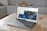 A sleek ASUS laptop is placed on a beige ottoman. The screen displays a webpage featuring a design with blue floral patterns. In the background, there is a grey couch with a patterned pillow and a wooden animal figurine.