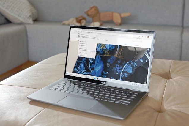 A sleek ASUS laptop is placed on a beige ottoman. The screen displays a webpage featuring a design with blue floral patterns. In the background, there is a grey couch with a patterned pillow and a wooden animal figurine.