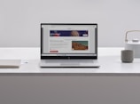 A laptop with an open webpage sits on a white marble table, surrounded by a notebook, a cup, and a kettle. The webpage shows a science-related article with images and text.