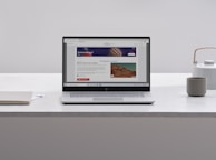 A laptop with an open webpage sits on a white marble table, surrounded by a notebook, a cup, and a kettle. The webpage shows a science-related article with images and text.