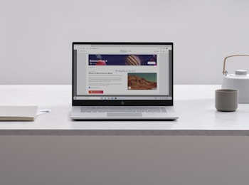A laptop with an open webpage sits on a white marble table, surrounded by a notebook, a cup, and a kettle. The webpage shows a science-related article with images and text.
