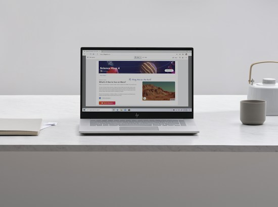 A laptop with an open webpage sits on a white marble table, surrounded by a notebook, a cup, and a kettle. The webpage shows a science-related article with images and text.