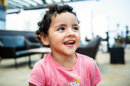 A young child with curly hair is sitting indoors and smiling joyfully, wearing a pink shirt featuring a unicorn. The background is blurred, showing some furniture like chairs and tables in a bright, airy space.