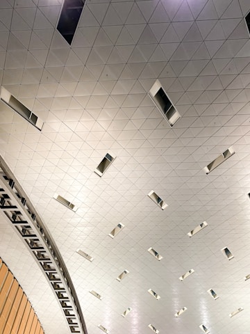 A modern, geometric ceiling design composed of triangular panels. The ceiling features recessed rectangular lights spaced evenly across the surface. In the foreground, a curved architectural element made of metal or wood adds dynamic visual interest.