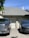 A residential setting features a gray brick house with a shingled roof and a chimney. In front of the garage, two vehicles are parked: a silver pickup truck on the left and a black SUV on the right. The house is adorned with some greenery, and the driveway has a shadow cast by nearby trees.