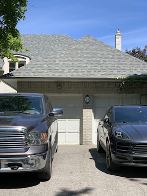 A residential setting features a gray brick house with a shingled roof and a chimney. In front of the garage, two vehicles are parked: a silver pickup truck on the left and a black SUV on the right. The house is adorned with some greenery, and the driveway has a shadow cast by nearby trees.