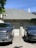 A residential setting features a gray brick house with a shingled roof and a chimney. In front of the garage, two vehicles are parked: a silver pickup truck on the left and a black SUV on the right. The house is adorned with some greenery, and the driveway has a shadow cast by nearby trees.