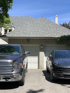 A residential setting features a gray brick house with a shingled roof and a chimney. In front of the garage, two vehicles are parked: a silver pickup truck on the left and a black SUV on the right. The house is adorned with some greenery, and the driveway has a shadow cast by nearby trees.