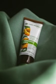 A tube of botanical scrub rests on a soft, green fabric. The packaging features an image of apricots and leaves, indicating it's a product with natural ingredients.