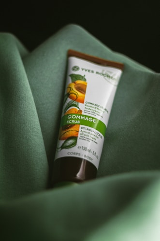 A tube of botanical scrub rests on a soft, green fabric. The packaging features an image of apricots and leaves, indicating it's a product with natural ingredients.