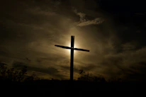 A cross silhouetted against a sunrise over a peaceful mountain path.