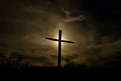 A cross silhouetted against a sunrise over a peaceful mountain path.