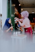 Two people are interacting at a small table outside, with one person pouring a drink while the other enjoys a dessert. They are both wearing hijabs, and the setting appears cozy and inviting with warm lighting and a casual atmosphere.