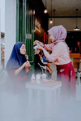 Two people are interacting at a small table outside, with one person pouring a drink while the other enjoys a dessert. They are both wearing hijabs, and the setting appears cozy and inviting with warm lighting and a casual atmosphere.