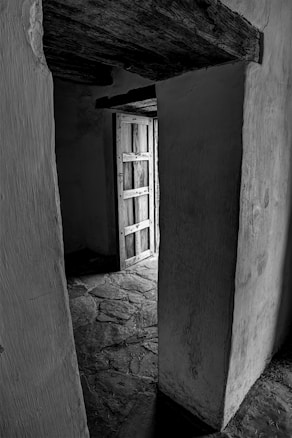A dimly lit corridor with a rough stone floor leading to an open wooden door. The walls and ceiling have a rustic texture, highlighted by the interplay of light and shadow.