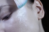 A close-up of a person's face focusing on the cheek area, with a soft-bristled brush applying white translucent powder. The skin texture is visible, showcasing pores and fine lines.