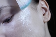 A close-up of a makeup brush applying soft pink blush on a cheek.