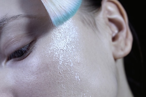 A close-up of a person's face focusing on the cheek area, with a soft-bristled brush applying white translucent powder. The skin texture is visible, showcasing pores and fine lines.