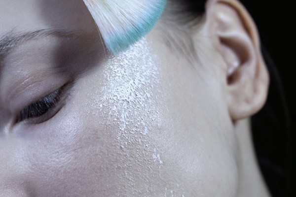 A close-up of a person's face focusing on the cheek area, with a soft-bristled brush applying white translucent powder. The skin texture is visible, showcasing pores and fine lines.