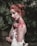 A woman with red hair is covered in blood, which stains her face, neck, shoulders, and hands. She is wearing a strapless, white garment with sequined details. Her hair is styled in an intricate updo, and she is standing against a lush, green, leafy background.