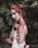 A woman with red hair is covered in blood, which stains her face, neck, shoulders, and hands. She is wearing a strapless, white garment with sequined details. Her hair is styled in an intricate updo, and she is standing against a lush, green, leafy background.