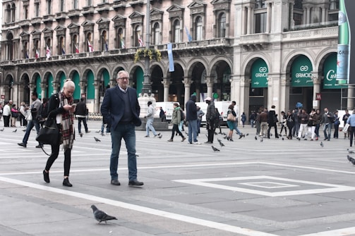 A busy urban square with people walking and interacting. The architecture in the background features a historic building with arched windows and numerous flags. Pigeons are scattered across the open, paved area. Two people are in the foreground; one is looking at a smartphone.
