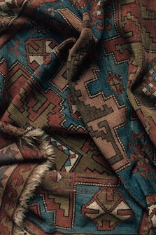 A richly patterned textile with intricate geometric designs featuring a mix of earthy tones like dark red, brown, and teal. The fabric appears to be woven and includes various symmetrical motifs that resemble traditional carpet designs. It has a slightly rough texture, accentuated by the presence of a fringe on one edge.