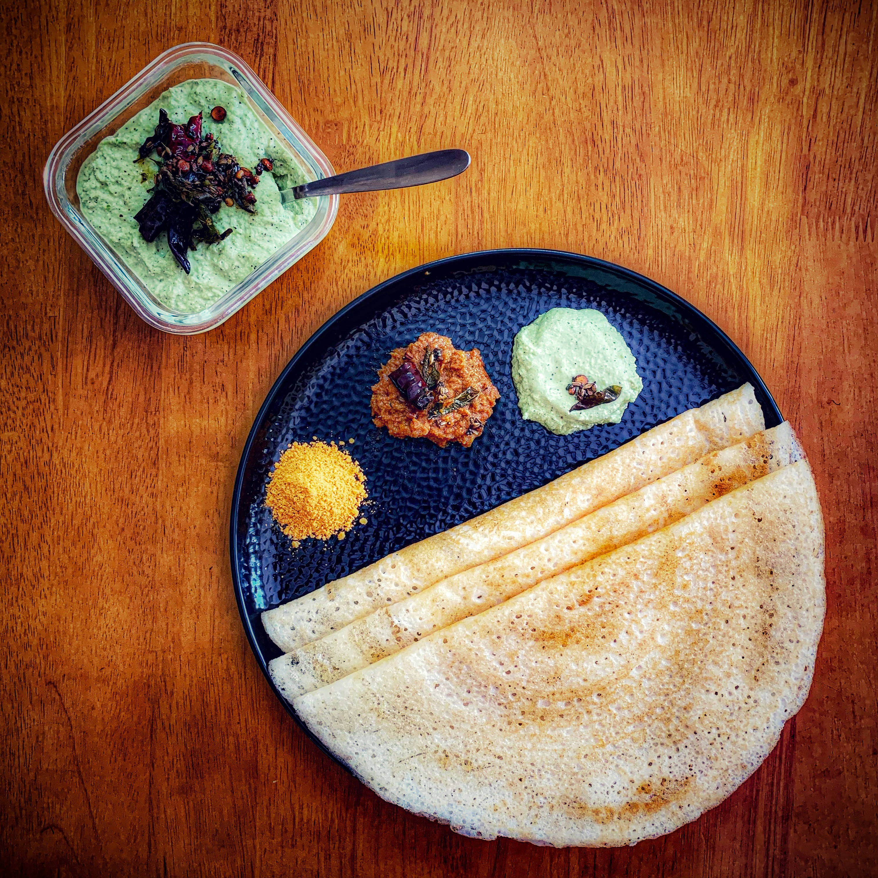 A plain dosa folded on a dark plate with podi, a red chutney and a coconut chutney