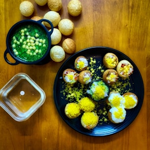 A wooden table displays a variety of small, crispy, round snacks filled with vibrant toppings. To the left, a black bowl contains a green, liquid-based dish with visible chickpeas. A black plate in the center holds several snacks garnished with yellow sev, white yogurt, and chopped onions, alongside a few scattered chickpeas. More of these spherical snacks are positioned around and on the plate, with a small transparent bowl nearby. The assortment suggests a colorful and flavorful culinary presentation.