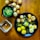 A wooden table displays a variety of small, crispy, round snacks filled with vibrant toppings. To the left, a black bowl contains a green, liquid-based dish with visible chickpeas. A black plate in the center holds several snacks garnished with yellow sev, white yogurt, and chopped onions, alongside a few scattered chickpeas. More of these spherical snacks are positioned around and on the plate, with a small transparent bowl nearby. The assortment suggests a colorful and flavorful culinary presentation.