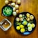 A wooden table displays a variety of small, crispy, round snacks filled with vibrant toppings. To the left, a black bowl contains a green, liquid-based dish with visible chickpeas. A black plate in the center holds several snacks garnished with yellow sev, white yogurt, and chopped onions, alongside a few scattered chickpeas. More of these spherical snacks are positioned around and on the plate, with a small transparent bowl nearby. The assortment suggests a colorful and flavorful culinary presentation.