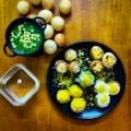 A wooden table displays a variety of small, crispy, round snacks filled with vibrant toppings. To the left, a black bowl contains a green, liquid-based dish with visible chickpeas. A black plate in the center holds several snacks garnished with yellow sev, white yogurt, and chopped onions, alongside a few scattered chickpeas. More of these spherical snacks are positioned around and on the plate, with a small transparent bowl nearby. The assortment suggests a colorful and flavorful culinary presentation.