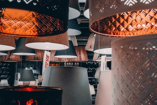 An assortment of various lampshades hanging in a store or showroom. The scene features a mix of textures and designs, including intricate geometric patterns and smooth surfaces. The lighting casts a warm glow, creating a cozy and inviting atmosphere.
