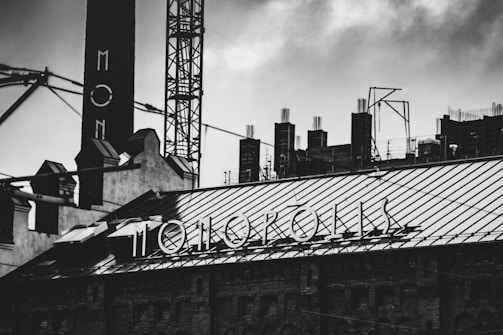 A black and white image of an industrial building. The rooftop features a metal framework with a prominent sign that reads 'MOTOKOPOLIS'. Various structural elements and scaffolding are visible in the background, suggesting construction or industrial activity.