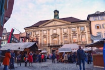 A bustling marketplace in front of a historic building with a clock tower. Several market stalls are set up, covered with fabric shades. People dressed in winter clothing are gathered around, engaging in conversations and browsing the stalls.
