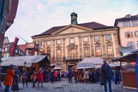 A bustling marketplace in front of a historic building with a clock tower. Several market stalls are set up, covered with fabric shades. People dressed in winter clothing are gathered around, engaging in conversations and browsing the stalls.