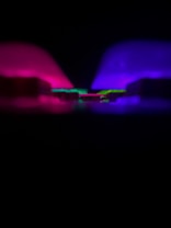 A dark setting is illuminated by vibrant neon lights. The left side features a deep magenta glow, while the right side displays a rich royal blue hue. In the center, a strip of bright green light creates a striking contrast against the darker surroundings.