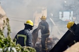 Firefighters wearing yellow helmets and protective gear are engaged in extinguishing a fire, surrounded by thick smoke and debris. They are actively using hoses and tools to control the fire in an outdoor setting, possibly in a residential area with visible greenery.
