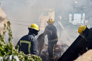 Firefighters wearing yellow helmets and protective gear are engaged in extinguishing a fire, surrounded by thick smoke and debris. They are actively using hoses and tools to control the fire in an outdoor setting, possibly in a residential area with visible greenery.