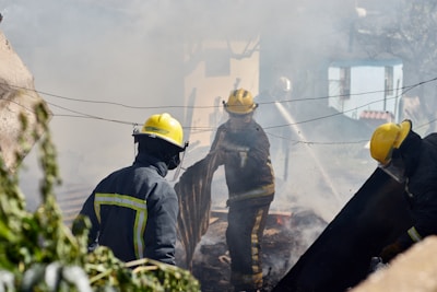 Firefighters wearing yellow helmets and protective gear are engaged in extinguishing a fire, surrounded by thick smoke and debris. They are actively using hoses and tools to control the fire in an outdoor setting, possibly in a residential area with visible greenery.