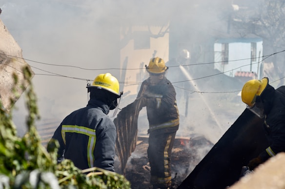 Firefighters wearing yellow helmets and protective gear are engaged in extinguishing a fire, surrounded by thick smoke and debris. They are actively using hoses and tools to control the fire in an outdoor setting, possibly in a residential area with visible greenery.