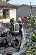 Two firefighters in protective gear spray water onto smoldering debris, suggesting they are working to extinguish a fire. The ground is covered with burned materials. Behind them, a partially damaged building appears charred around the windows. A group of people stands in the background, some wearing bright clothing, observing the scene. Nearby greenery is slightly blurred, framing the image.