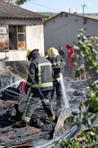 Two firefighters in protective gear spray water onto smoldering debris, suggesting they are working to extinguish a fire. The ground is covered with burned materials. Behind them, a partially damaged building appears charred around the windows. A group of people stands in the background, some wearing bright clothing, observing the scene. Nearby greenery is slightly blurred, framing the image.