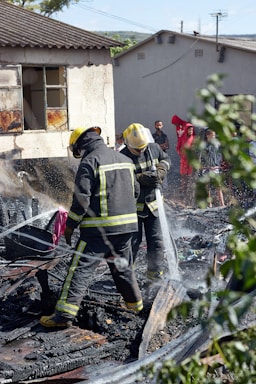 Two firefighters in protective gear spray water onto smoldering debris, suggesting they are working to extinguish a fire. The ground is covered with burned materials. Behind them, a partially damaged building appears charred around the windows. A group of people stands in the background, some wearing bright clothing, observing the scene. Nearby greenery is slightly blurred, framing the image.
