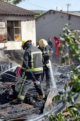 Two firefighters in protective gear spray water onto smoldering debris, suggesting they are working to extinguish a fire. The ground is covered with burned materials. Behind them, a partially damaged building appears charred around the windows. A group of people stands in the background, some wearing bright clothing, observing the scene. Nearby greenery is slightly blurred, framing the image.