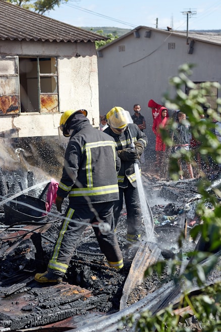 Two firefighters in protective gear spray water onto smoldering debris, suggesting they are working to extinguish a fire. The ground is covered with burned materials. Behind them, a partially damaged building appears charred around the windows. A group of people stands in the background, some wearing bright clothing, observing the scene. Nearby greenery is slightly blurred, framing the image.