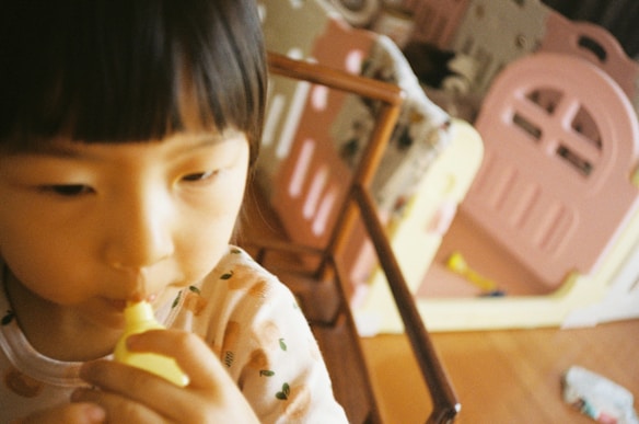 A young child with black hair is holding a yellow object close to their mouth. They are wearing a light-colored shirt with small patterns. In the background, there is a pink playpen or play area, and the floor is wooden with some scattered toys.