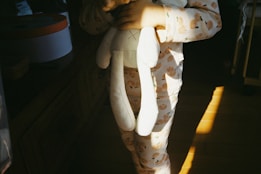 A child wearing pajamas with a peach pattern is holding a white stuffed toy bunny. Warm sunlight creates a play of shadows on the floor, adding a cozy atmosphere to the scene.